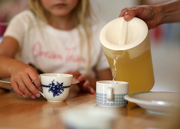 Ein Kind sitzt am Tisch und bekommt einen Tee eingeschenkt