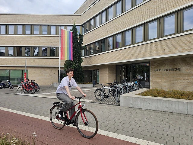 Ein junger Mensch sitzt vor dem Haus der Kirche Niendorf auf einem Fahrrad